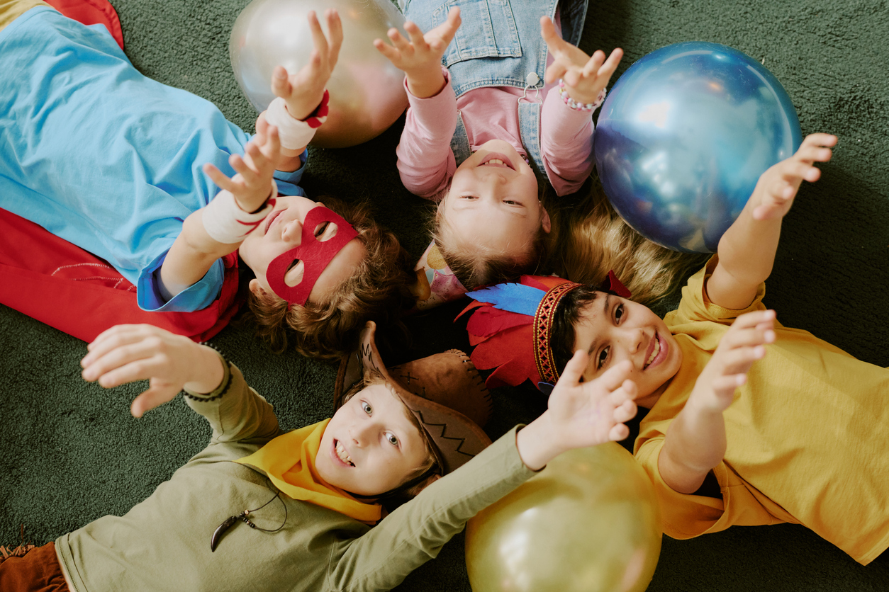 Group of multiethnic children lying on floor reaching hands toward camera smiling, wearing costumes and playing with balloons during playful indoor activity